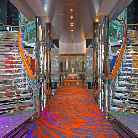 Golden Sands Boutique Hotel-Dubai Creek Εσωτερικό φωτογραφία The photo shows a modern, elegantly designed interior space, likely on a cruise ship. It features two sweeping, curved staircases made of glass and metal, flanking either side of a central corridor. The ground is covered with a vibrant orange carpet 
