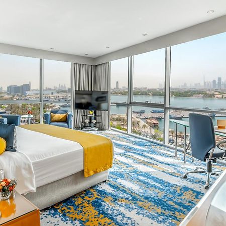Golden Sands Boutique Hotel-Dubai Creek Εξωτερικό φωτογραφία The photo shows a modern hotel room with a spacious layout and large windows that offer a view of the city and water. The room features a comfortable bed with a bright yellow and white duvet, a stylish area rug with a blue and yellow pattern, and con