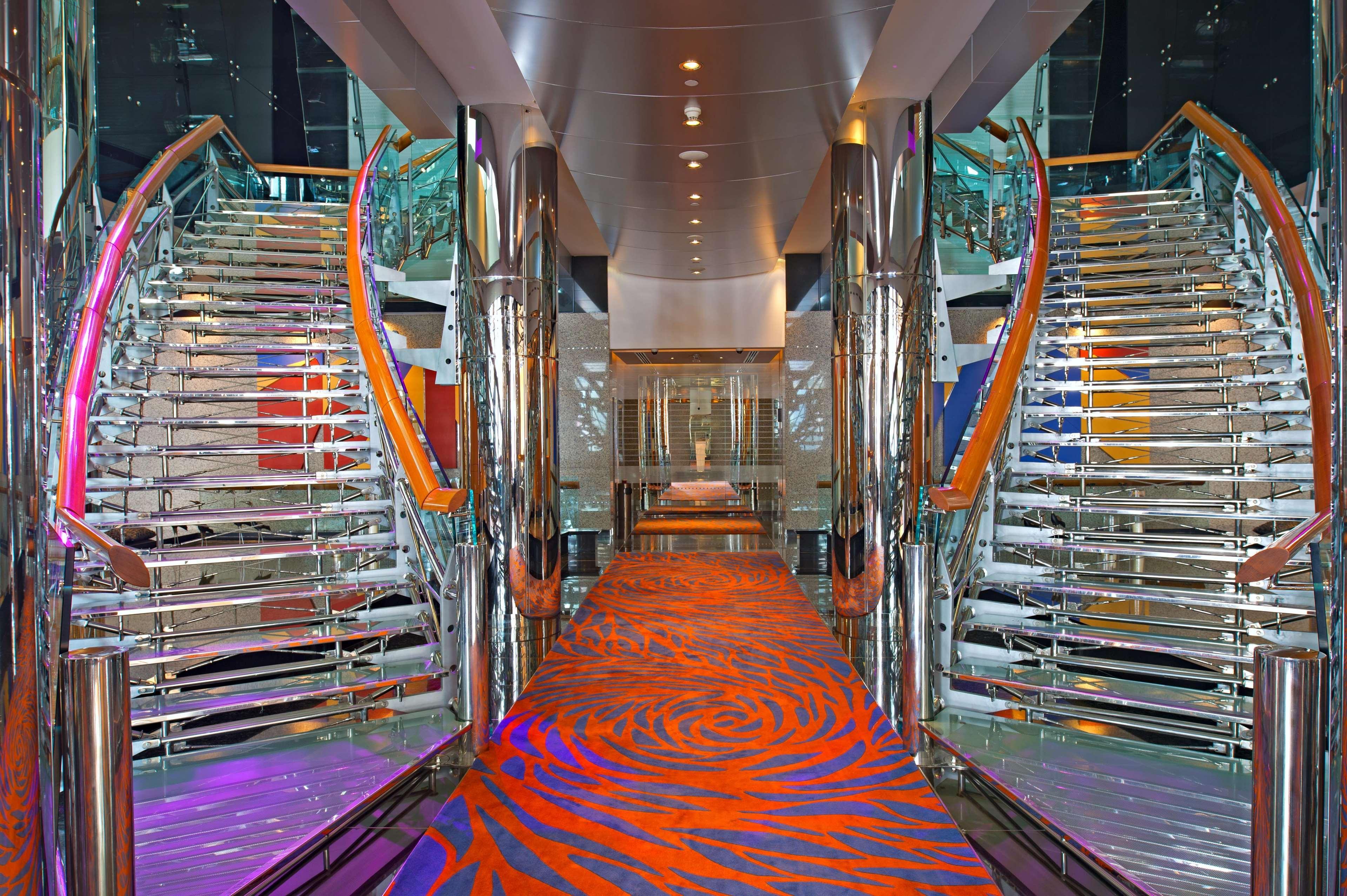 Golden Sands Boutique Hotel-Dubai Creek Εσωτερικό φωτογραφία The photo shows a modern, elegantly designed interior space, likely on a cruise ship. It features two sweeping, curved staircases made of glass and metal, flanking either side of a central corridor. The ground is covered with a vibrant orange carpet 