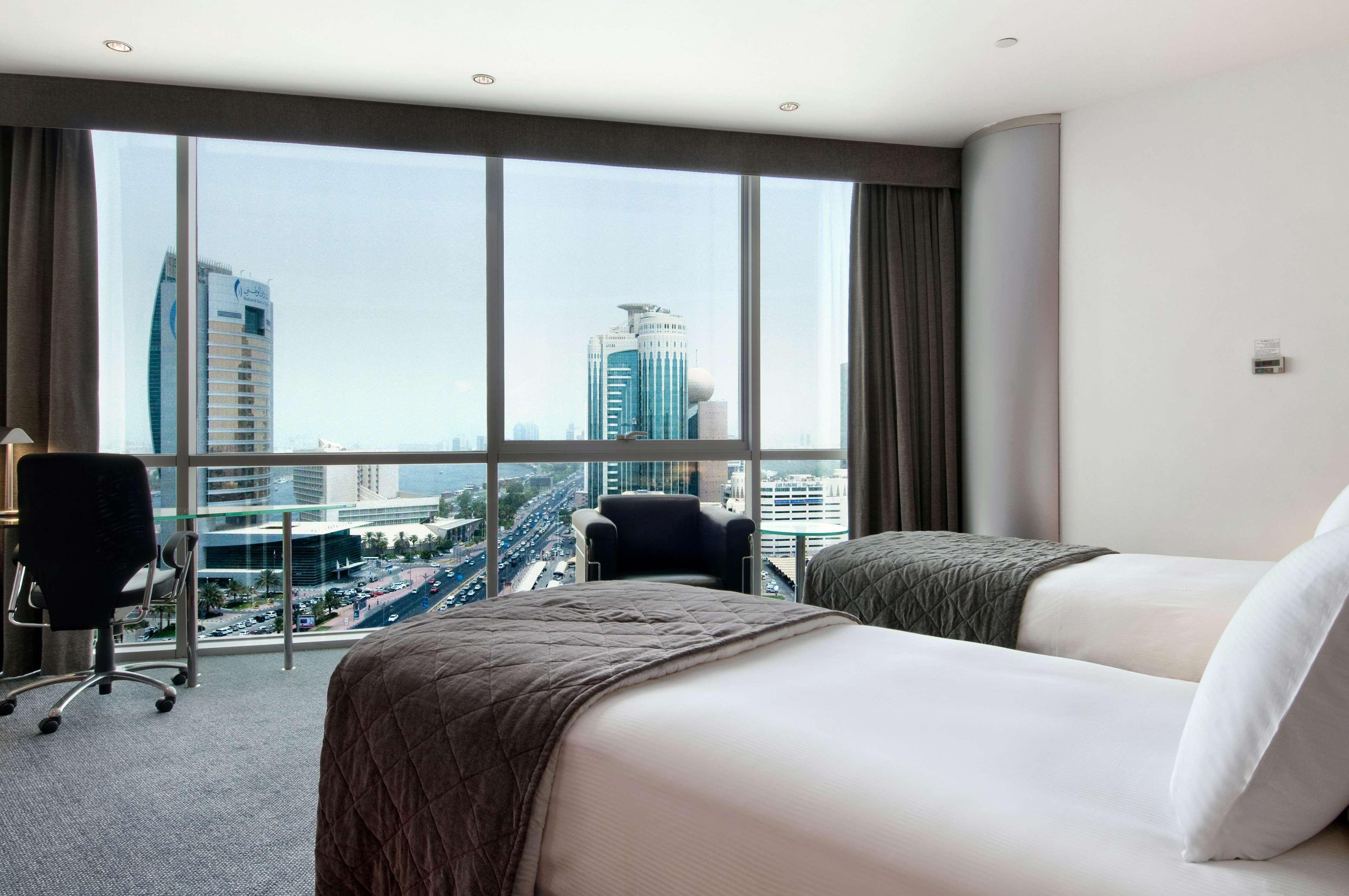 Golden Sands Boutique Hotel-Dubai Creek Δωμάτιο φωτογραφία The photo shows a modern hotel room with two beds. The beds are neatly made with white linens and dark gray throws. There is a large window that offers a view of a cityscape, featuring tall buildings and skyscrapers. The room has contemporary furnish
