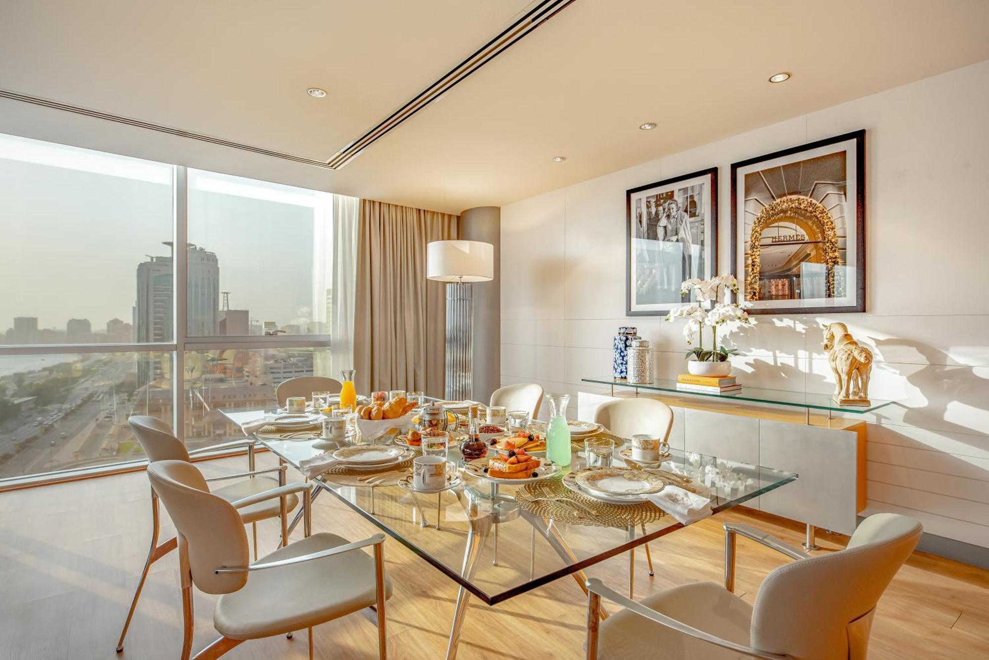 Golden Sands Boutique Hotel-Dubai Creek Δωμάτιο φωτογραφία The photo shows a modern dining room featuring a glass dining table set for a meal. The table is adorned with plates, cutlery, glasses, and a variety of food items, suggesting a breakfast or brunch setting. Surrounding the table are sleek, white chai
