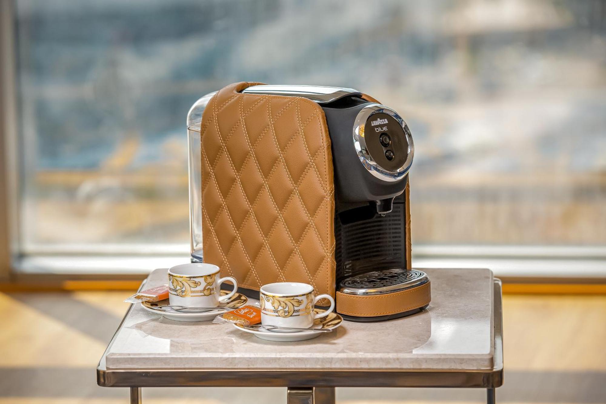 Golden Sands Boutique Hotel-Dubai Creek Εξωτερικό φωτογραφία The photo shows a stylish coffee machine with a quilted brown cover, placed on a table. Beside it are two elegant cups of coffee, each accompanied by small plates with a biscuit or cookie. The background appears to have a bright, airy setting, possib