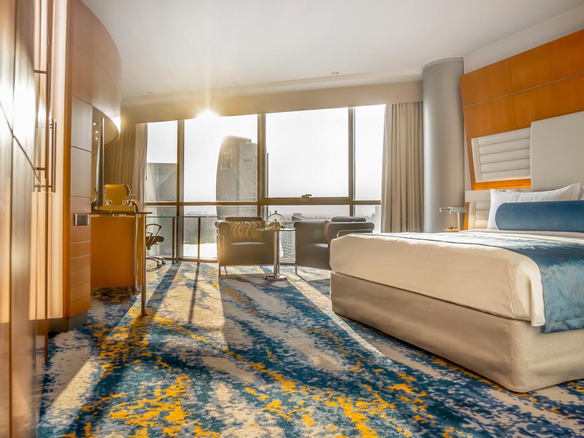 Golden Sands Boutique Hotel-Dubai Creek Δωμάτιο φωτογραφία The photo shows a modern hotel room featuring a large bed with a light beige cover and a blue accent blanket. Large windows allow natural light to stream in, revealing a scenic view outside. The floor is adorned with a colorful area rug in shades of 