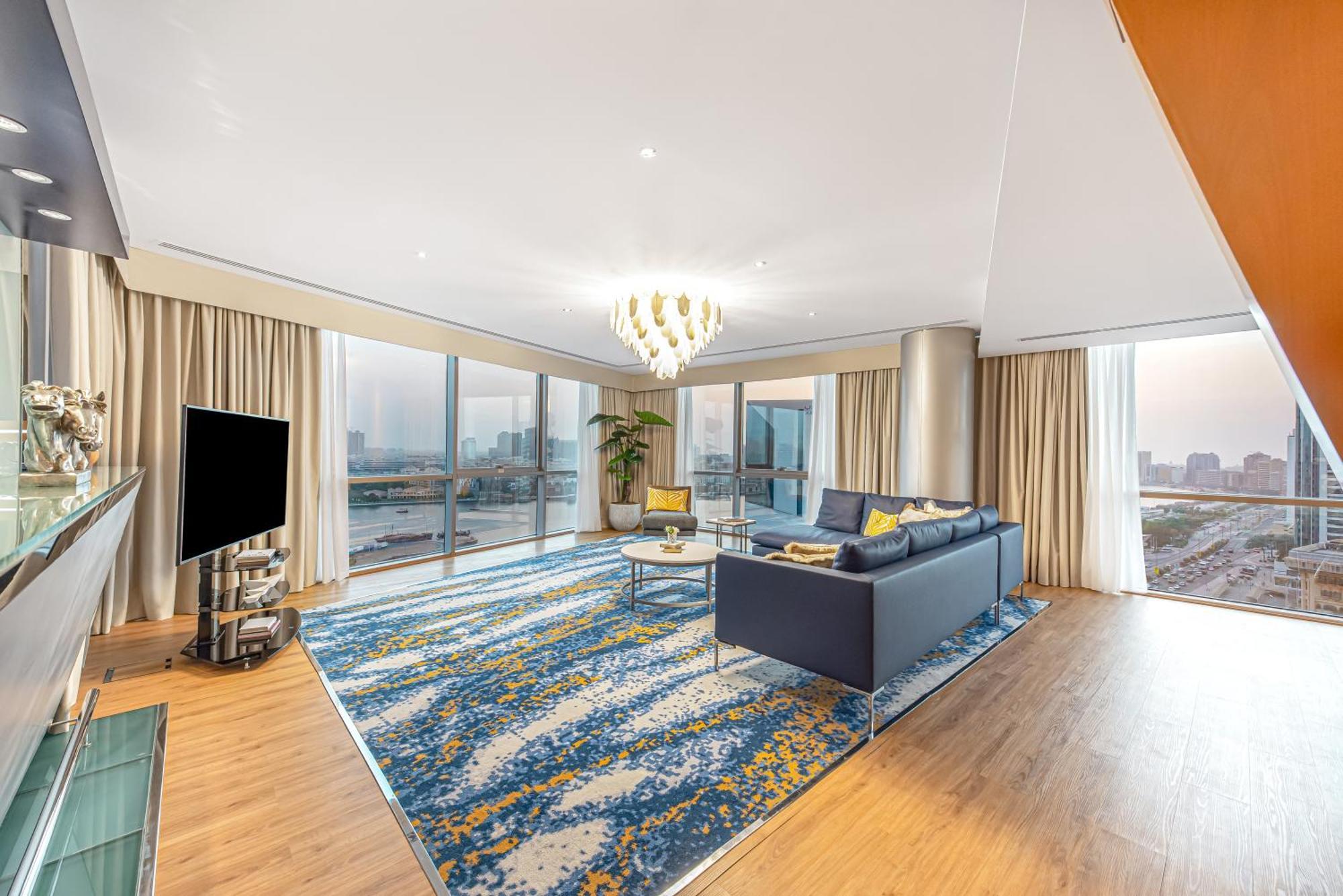 Golden Sands Boutique Hotel-Dubai Creek Δωμάτιο φωτογραφία The photo shows a spacious and modern living room with large windows that provide a view of an urban landscape. The room features a stylish blue sofa with decorative pillows, a coffee table, and a vibrant area rug with a colorful pattern. A sleek tel
