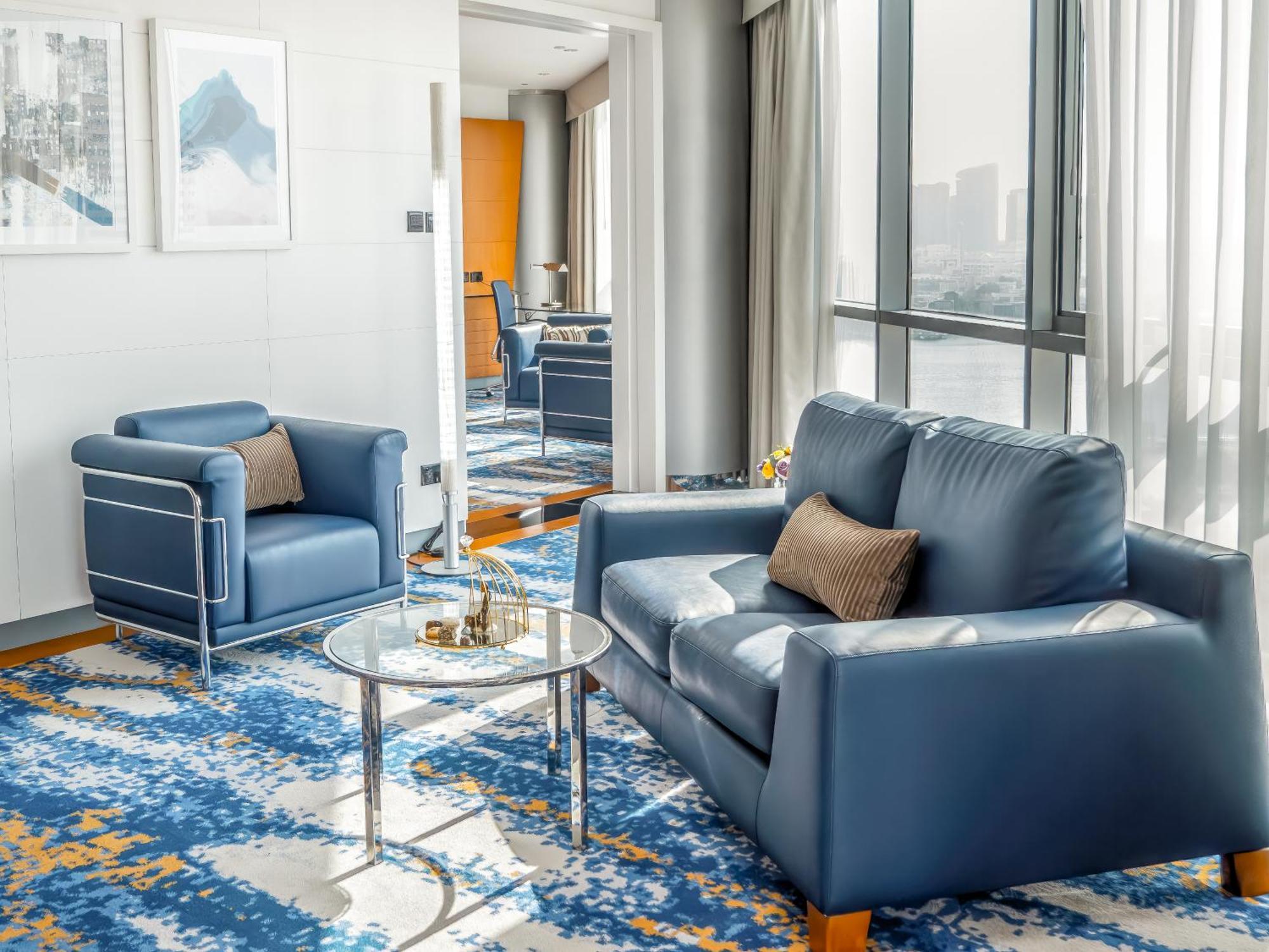 Golden Sands Boutique Hotel-Dubai Creek Δωμάτιο φωτογραφία The photo shows a stylish, modern living area with two pieces of furniture. There is a blue leather sofa with a brown cushion placed on it, accompanied by a matching blue armchair. In front of the sofa, there is a glass coffee table. The floor has a 