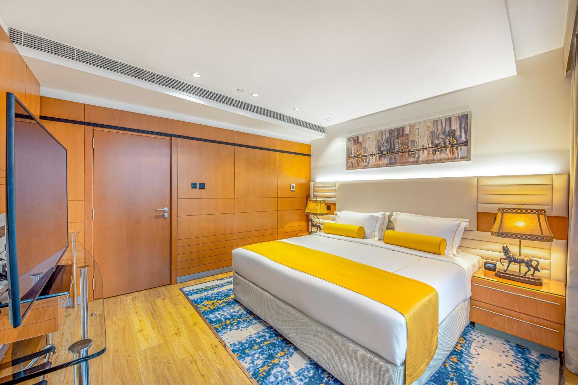 Golden Sands Boutique Hotel-Dubai Creek Δωμάτιο φωτογραφία The photo shows a modern hotel room featuring a king-sized bed with bright yellow accents on the bedding. The walls are a warm wood tone, with a large piece of artwork above the bed. There are nightstands on either side of the bed, each with lamps. A