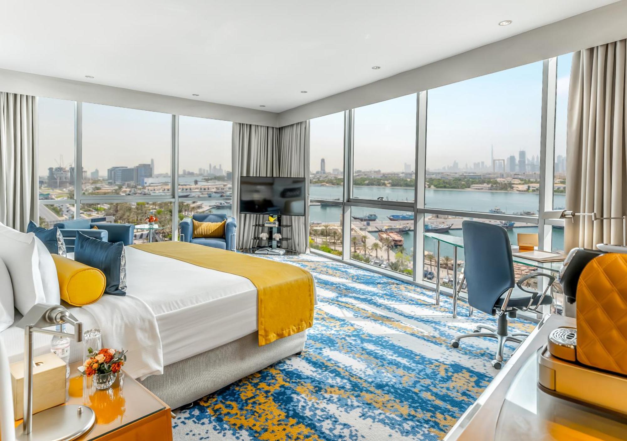 Golden Sands Boutique Hotel-Dubai Creek Εξωτερικό φωτογραφία The photo shows a modern hotel room with a spacious layout and large windows that offer a view of the city and water. The room features a comfortable bed with a bright yellow and white duvet, a stylish area rug with a blue and yellow pattern, and con