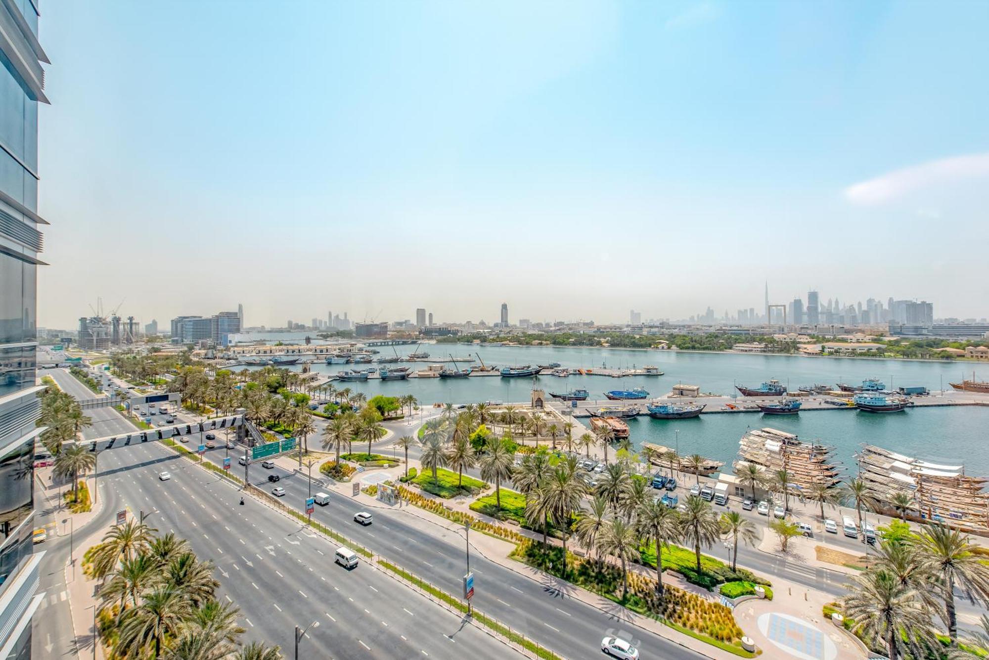 Golden Sands Boutique Hotel-Dubai Creek Εξωτερικό φωτογραφία The photo showcases a scenic view of a city waterfront. In the foreground, there is a wide road lined with palms, indicating a tropical or subtropical climate. Vehicles can be seen traveling along the road. 

Across the water, there are several boats
