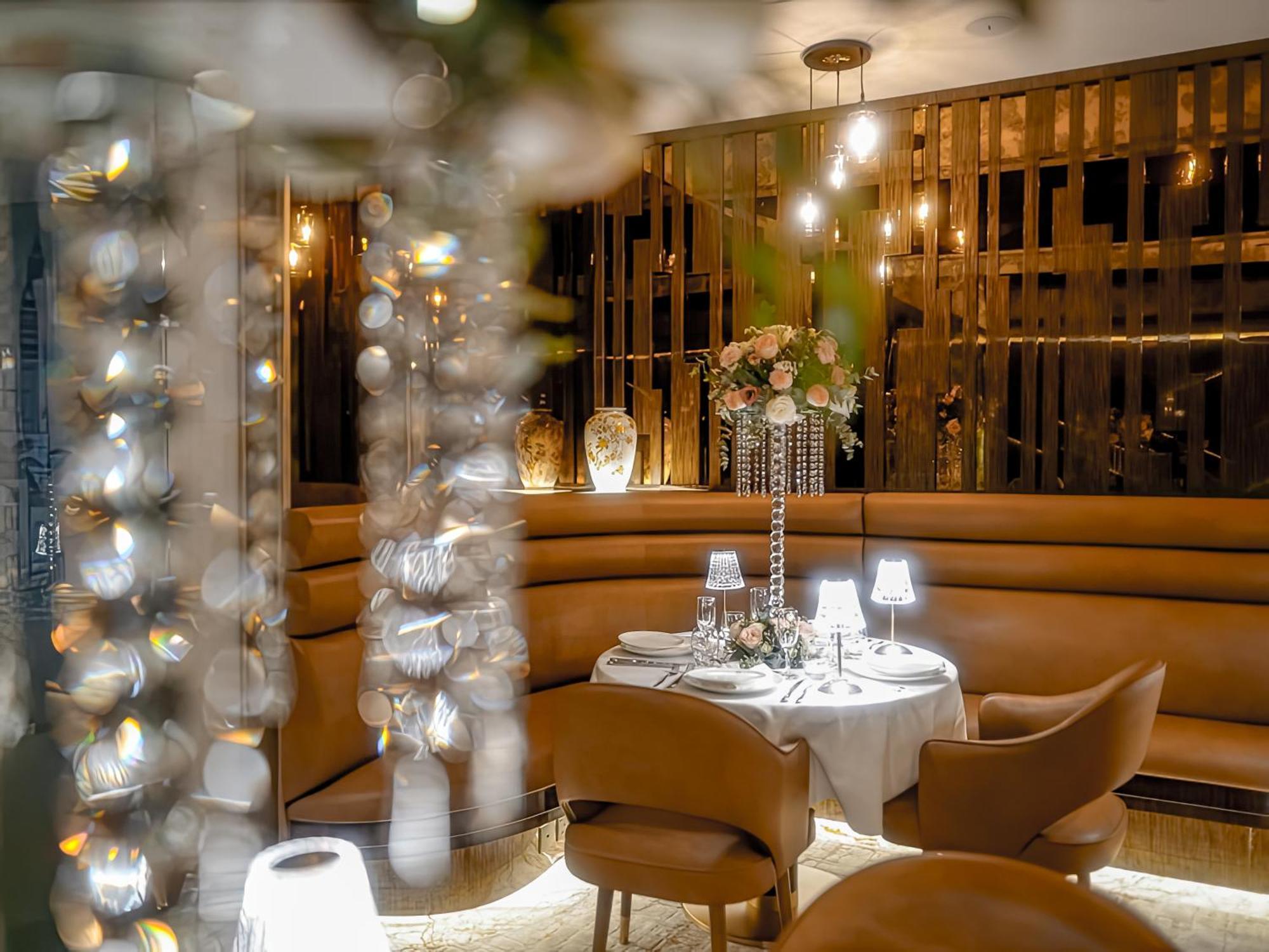 Golden Sands Boutique Hotel-Dubai Creek Εξωτερικό φωτογραφία The photo depicts an elegant restaurant interior. There is a round dining table set for two, complete with a white tablecloth, center floral arrangement, and small lamps. The seating consists of curved, brown leather chairs, suggesting a cozy atmosph