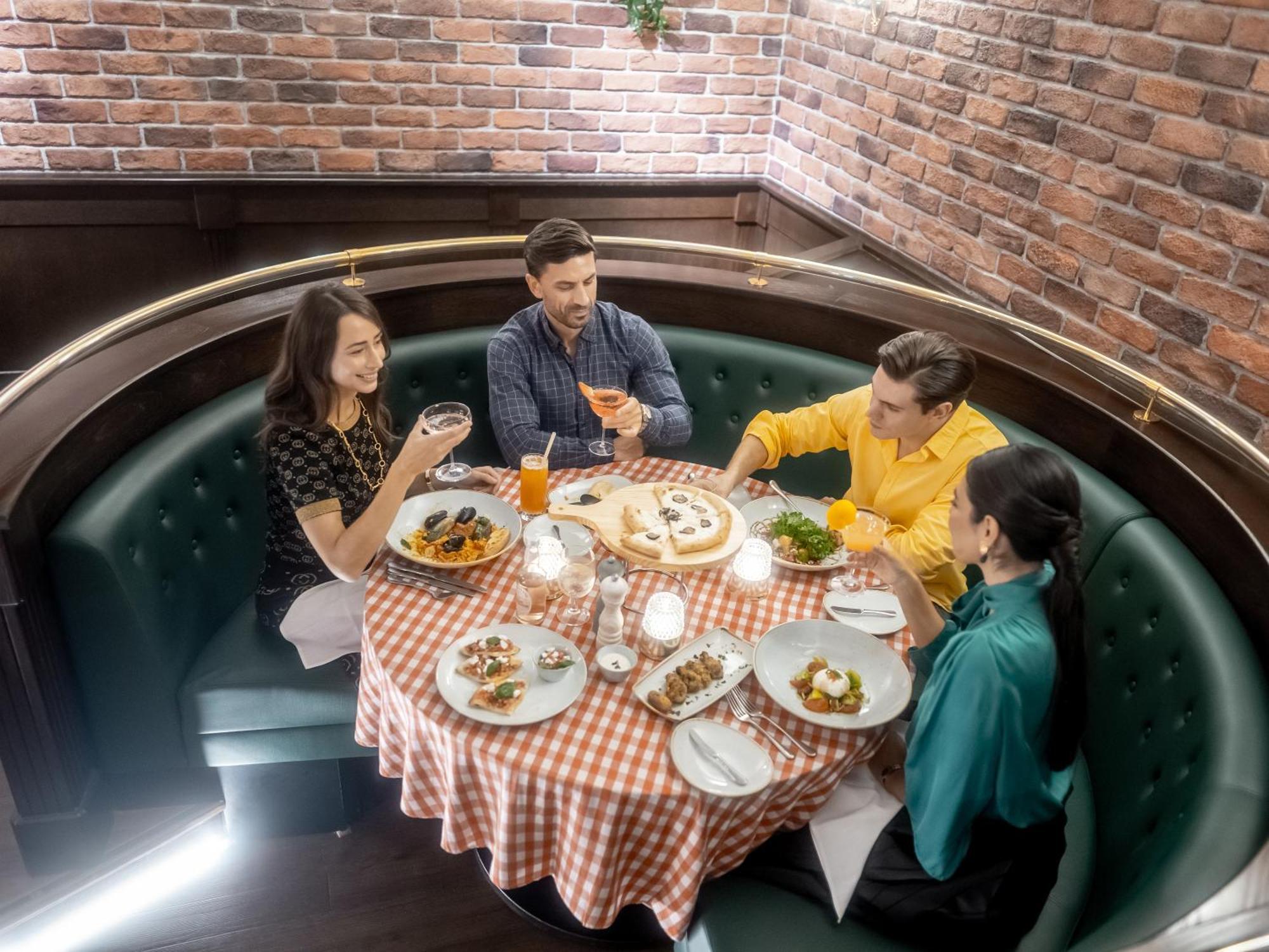 Golden Sands Boutique Hotel-Dubai Creek Εξωτερικό φωτογραφία The photo shows a cozy dining setting featuring four people sitting around a round table. The table is covered with a checkered tablecloth and is surrounded by a cushioned booth. There are several plates of food displayed, and each person has a drink