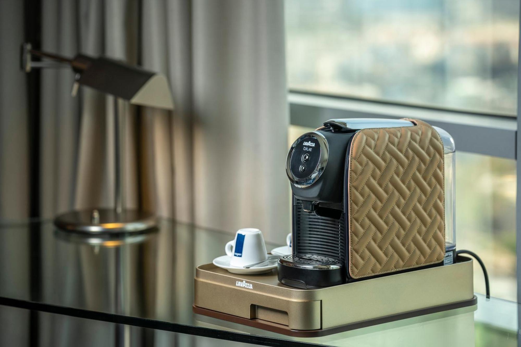 Golden Sands Boutique Hotel-Dubai Creek Δωμάτιο φωτογραφία The photo features a coffee machine placed on a sleek table. The machine has a stylish design with a patterned brown exterior. Next to the machine, there is a small saucer with a blue and white coffee cup. The background includes a large window with 