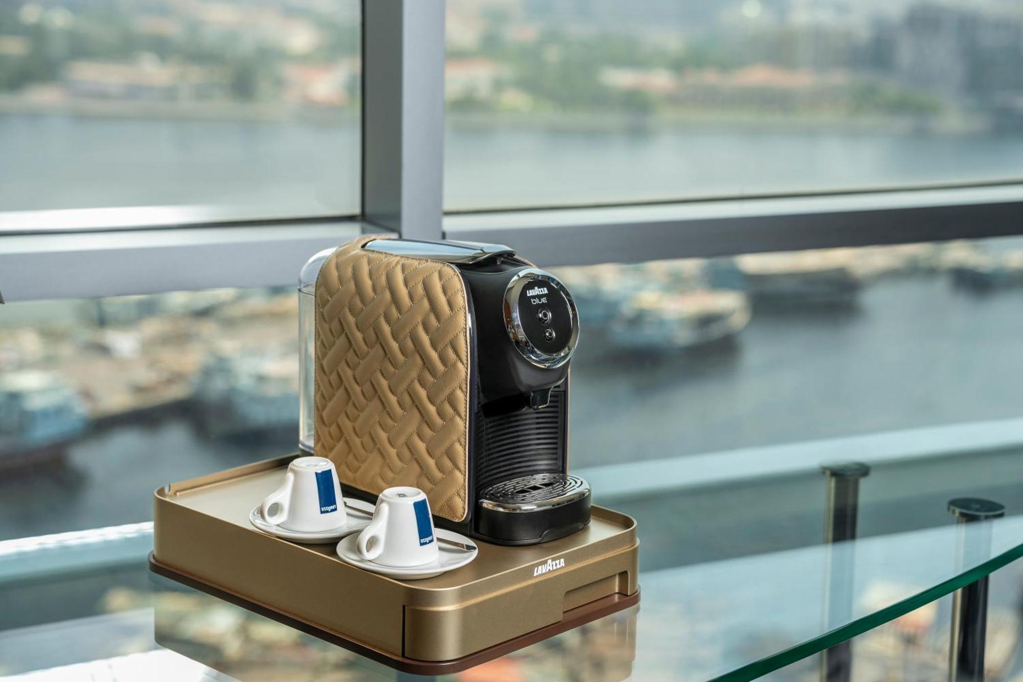 Golden Sands Boutique Hotel-Dubai Creek Εξωτερικό φωτογραφία The image shows a coffee machine placed on a glass table with a scenic view of water in the background. The coffee machine has a sleek design, featuring a beige and black color scheme. There are two coffee cups positioned next to the machine, along w